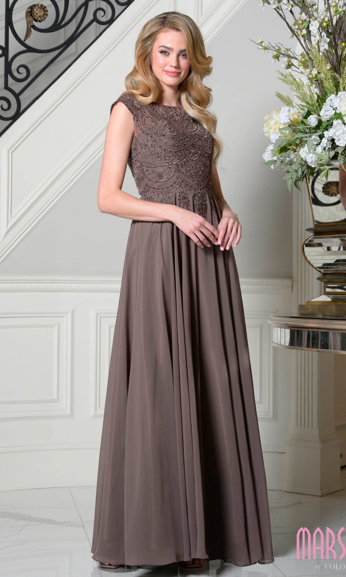 A woman poses indoors near a floral display and decorative staircase, wearing the Marsoni M238 long formal dress in dark taupe—a sleeveless, floor-length gown with an embellished lace bodice and flowing skirt.